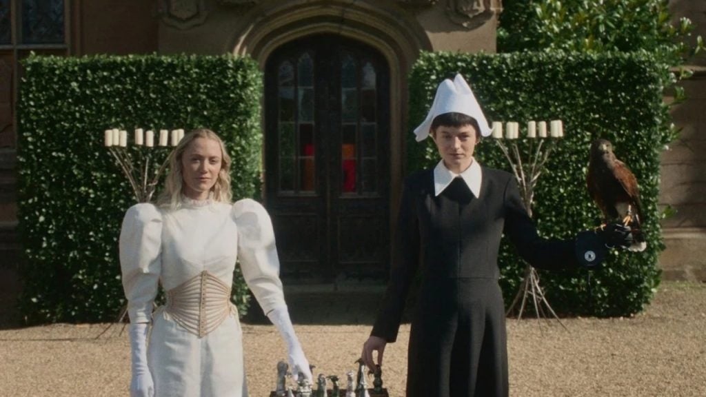 Two women stand outdoors in front of a building, one in a white dress and the other in black holding a bird of prey; a chessboard is set on the table between them.