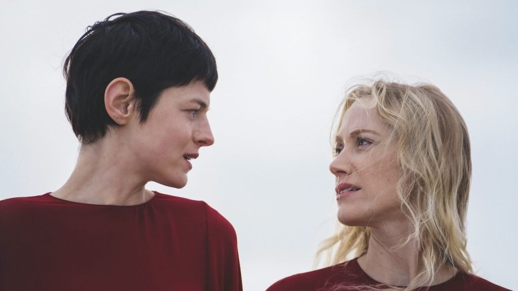 Two women wearing red tops stand close together outdoors, facing each other and making eye contact against a pale sky background.