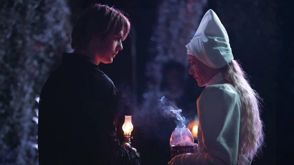 A man and a woman in historical costumes hold lanterns at night; the woman wears a tall white headdress and smoke rises from her lantern.
