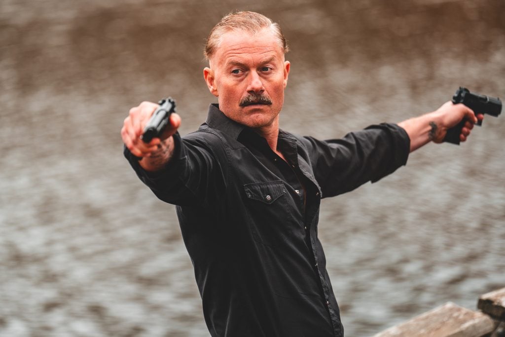 A man with a mustache in a black shirt stands by water, holding handguns in both hands and pointing them in opposite directions.