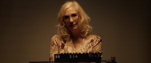 An older woman with long blonde hair and glasses stands behind a mugshot board reading "BK 234 1963" in a dimly lit room.