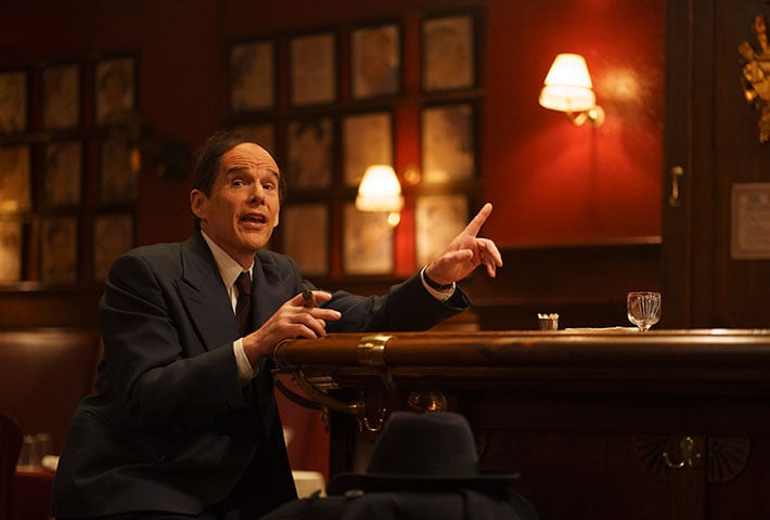 A man in a suit sits at a bar, gesturing with one hand while holding a cigarette, with framed pictures and warm lighting in the background.
