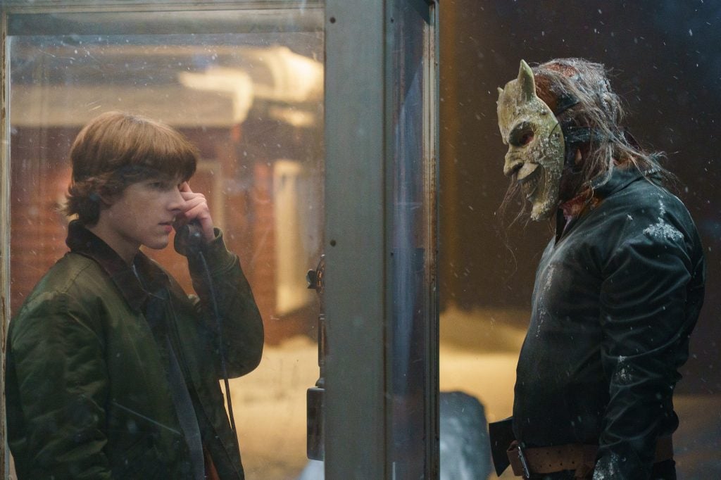 A boy stands inside a phone booth using the phone, while a person wearing a creepy mask and dark clothing stands outside, looking at him. Snow is falling around them.