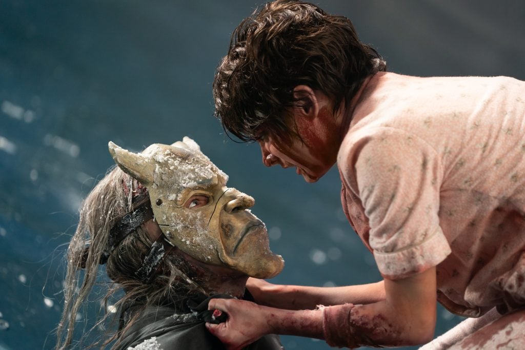 A person in a bloody shirt confronts another person wearing a horned mask and covered in dirt, gripping their collar in an intense scene.