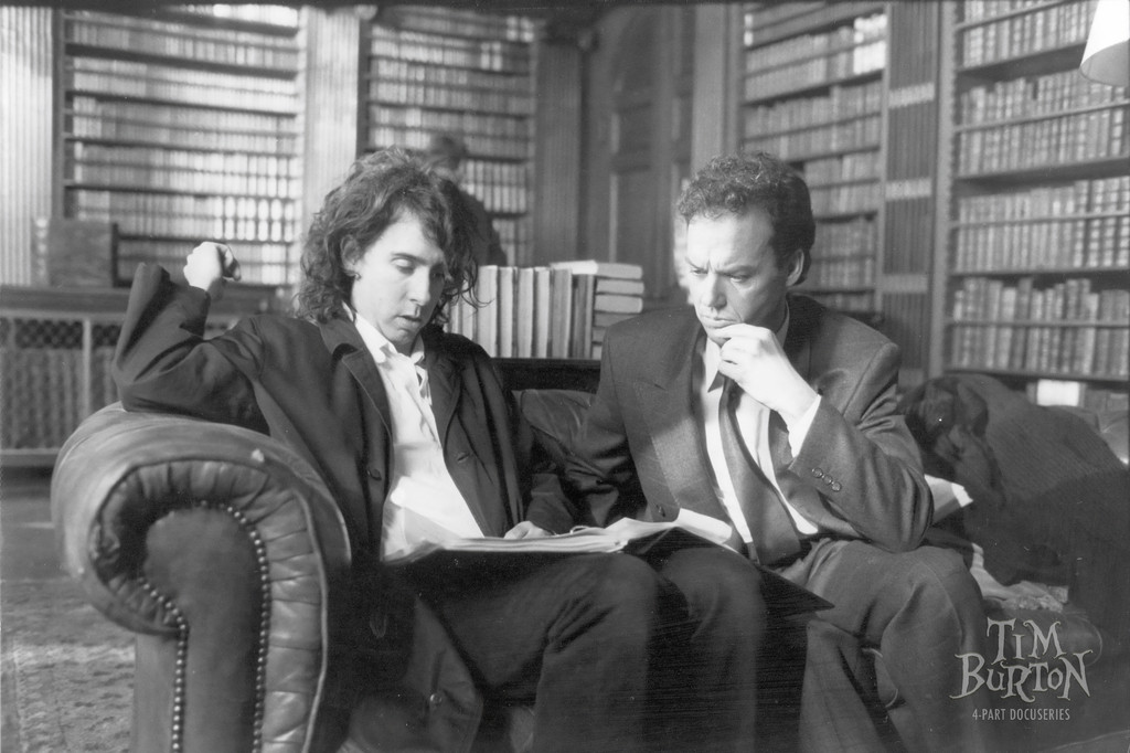Two men in suits sit on a couch in a library, looking at a book together. Shelves of books line the background. "Tim Burton 4-Part Docuseries" text is visible in the corner.