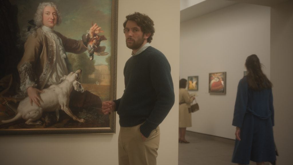 A man stands next to a large framed painting in an art gallery, with other visitors and artworks visible in the background.