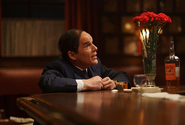 A man in a suit sits at a bar next to a bottle of whiskey, a glass, and a vase of red flowers, looking up and appearing to speak.