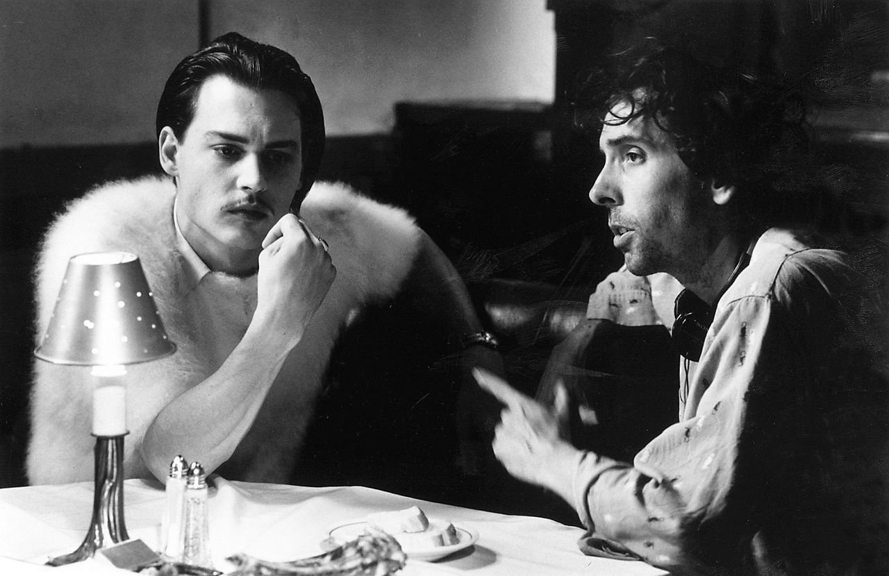 Two men sit at a restaurant table engaged in conversation, with a lamp and table settings visible in front of them.