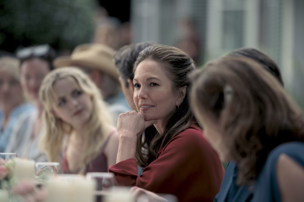 A woman in a red blouse sits at a table outdoors among a group of people, looking ahead with her hand resting on her chin.