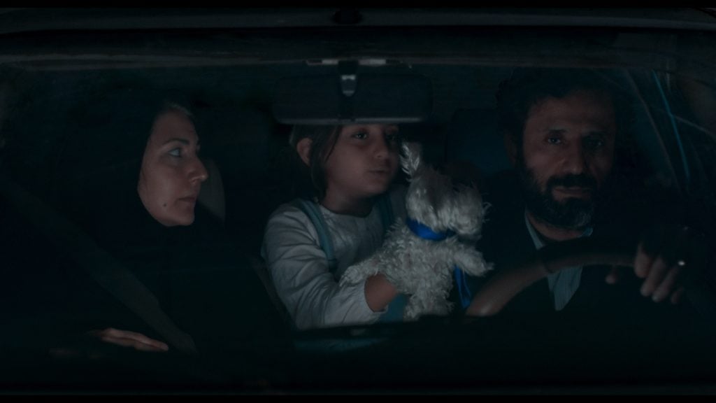 Three people sit in a car at night; a woman on the left, a child holding a toy dog in the middle, and a man driving on the right.