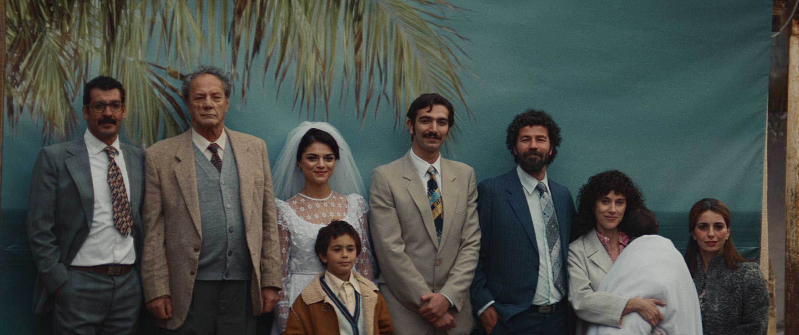 A group of people dressed formally pose for a photo in front of a backdrop with palm leaves; one woman wears a wedding dress, another holds a baby, and a child stands in front.