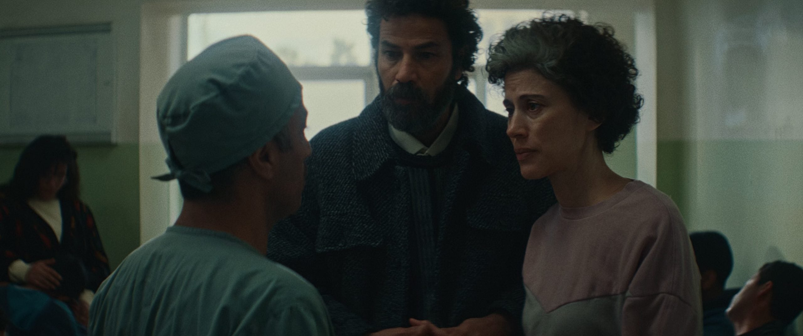 A doctor in scrubs speaks with a man and a woman in a hospital waiting room, with other people seated in the background.