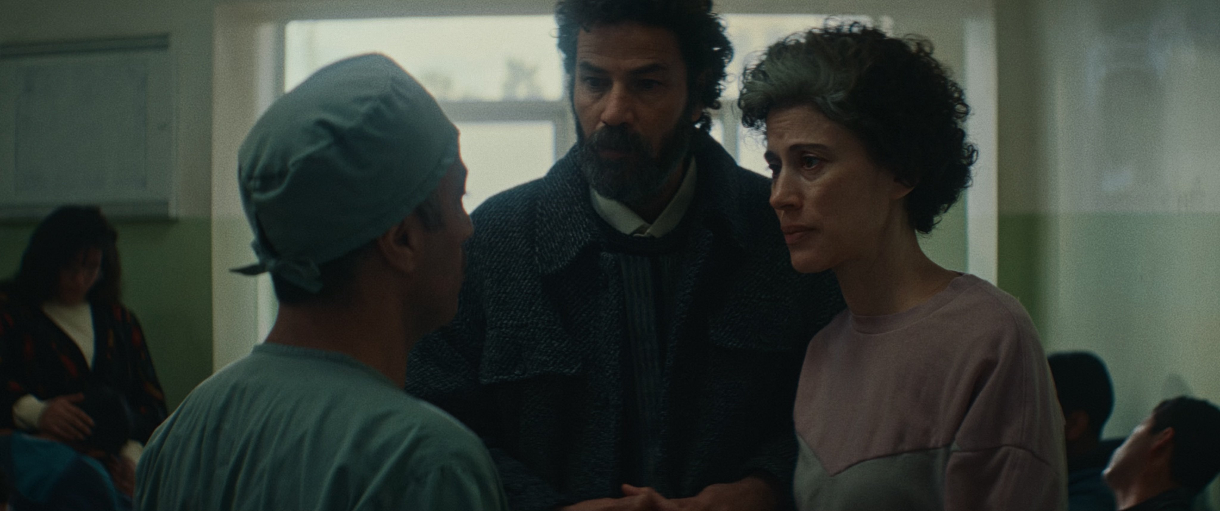 A doctor in scrubs speaks with a man and a woman in a hospital waiting room, with other people seated in the background.