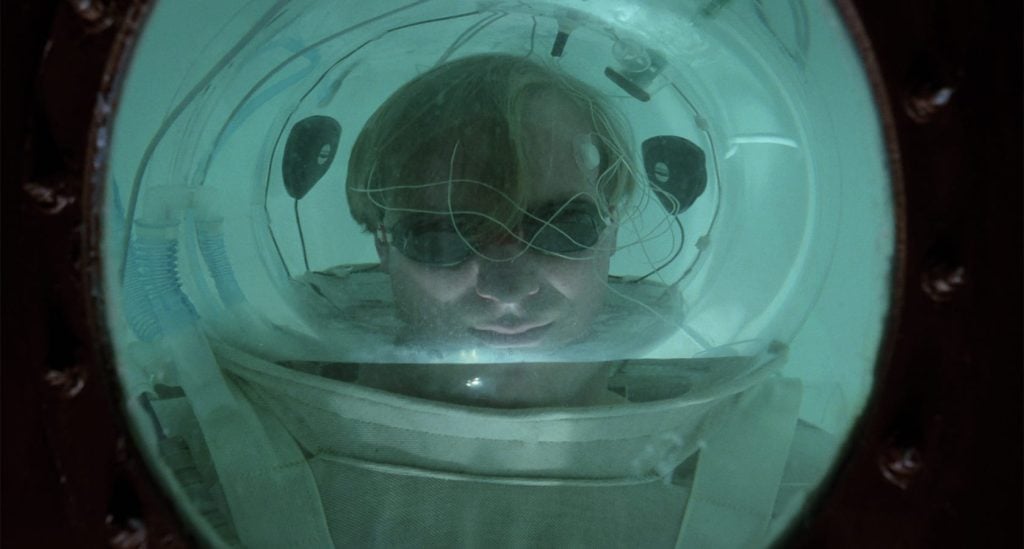 A person wearing goggles and headgear is viewed through a round glass window, submerged in water inside a confined space or vessel.