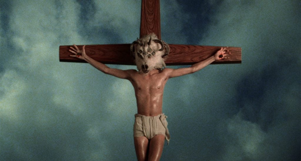 A person wearing a horned animal skull mask and loincloth is depicted crucified on a wooden cross against a cloudy sky background.