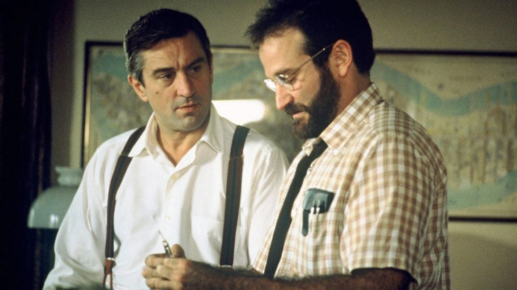 Two men stand indoors; one wears a white shirt with suspenders, while the other wears glasses and a checkered shirt, holding a pen and paper. They appear to be having a serious conversation.