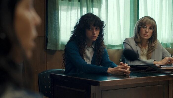 Two women sit at a table in an office or interrogation room, attentively listening to a person speaking out of focus in the foreground.