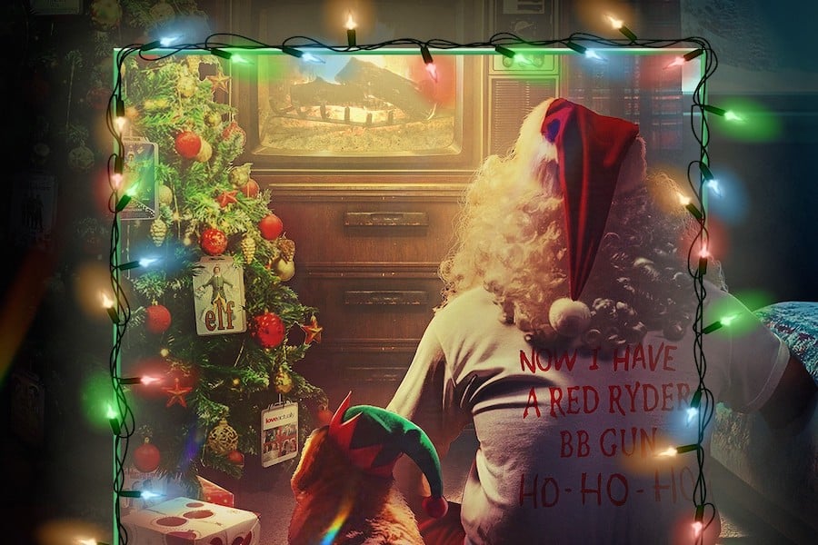 Person with curly hair and a cat, both wearing festive outfits, sit near a decorated Christmas tree watching a TV; string lights frame the scene.