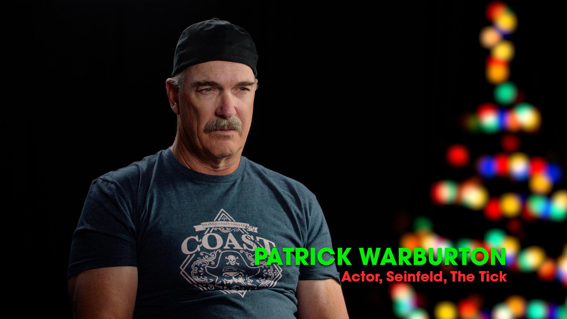 A man in a dark t-shirt and backwards cap sits in front of a black background with blurred colorful lights and text reading "Patrick Warburton, Actor, Seinfeld, The Tick.