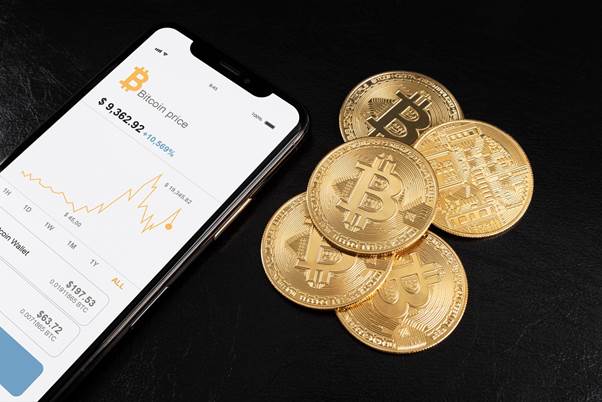A smartphone displaying a Bitcoin price chart sits next to five physical Bitcoin coins on a black surface.
