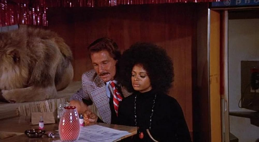 A man in a plaid suit and a woman with an afro sit at a table with papers and a red lamp; a lion lies on a counter behind them.