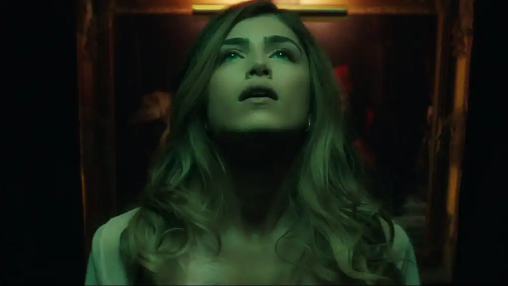A woman with long blonde hair looks upward with her mouth slightly open, illuminated by moody, greenish lighting in a dark setting.
