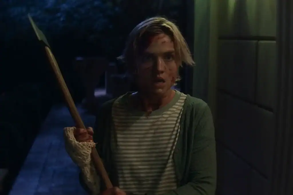 A woman with short blonde hair, wearing a striped shirt and green cardigan, stands outside at night holding a spear. Her face is bloodied and she looks alarmed.