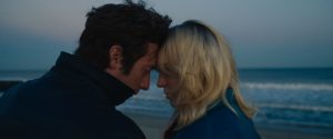 A man and woman stand close together on a beach at dusk, touching foreheads and looking down.