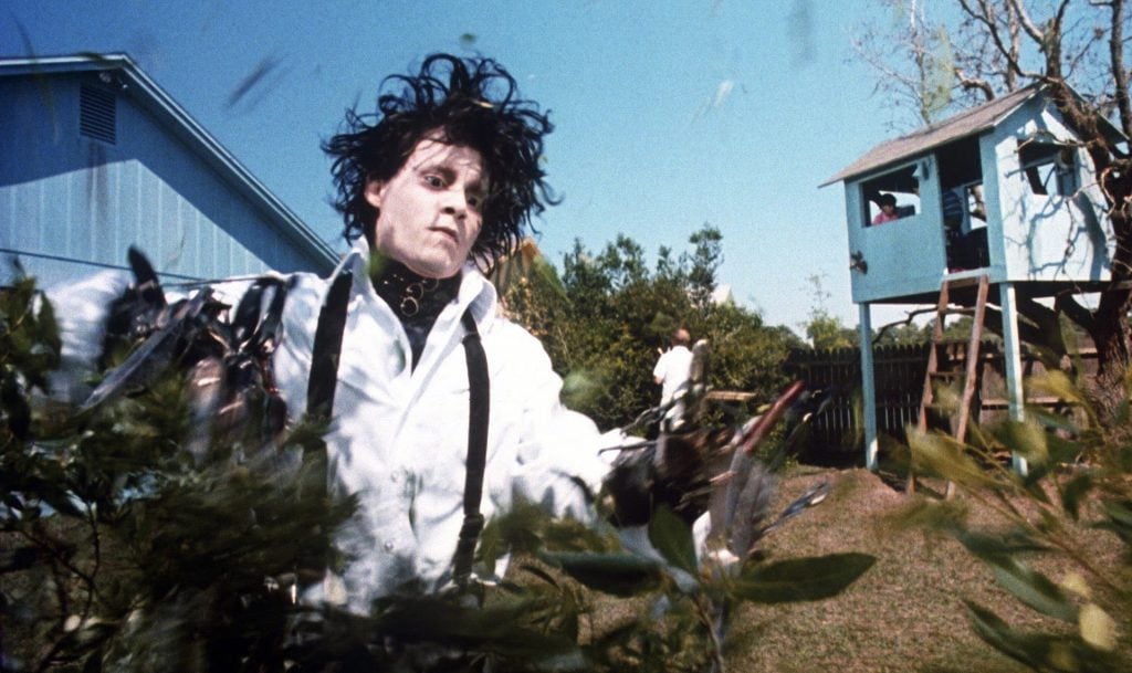 A pale man with wild black hair and scissor hands trims bushes in a suburban yard with a blue house and a wooden treehouse in the background.