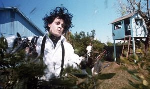 A pale man with wild black hair and scissor hands trims bushes in a suburban yard with a blue house and a wooden treehouse in the background.