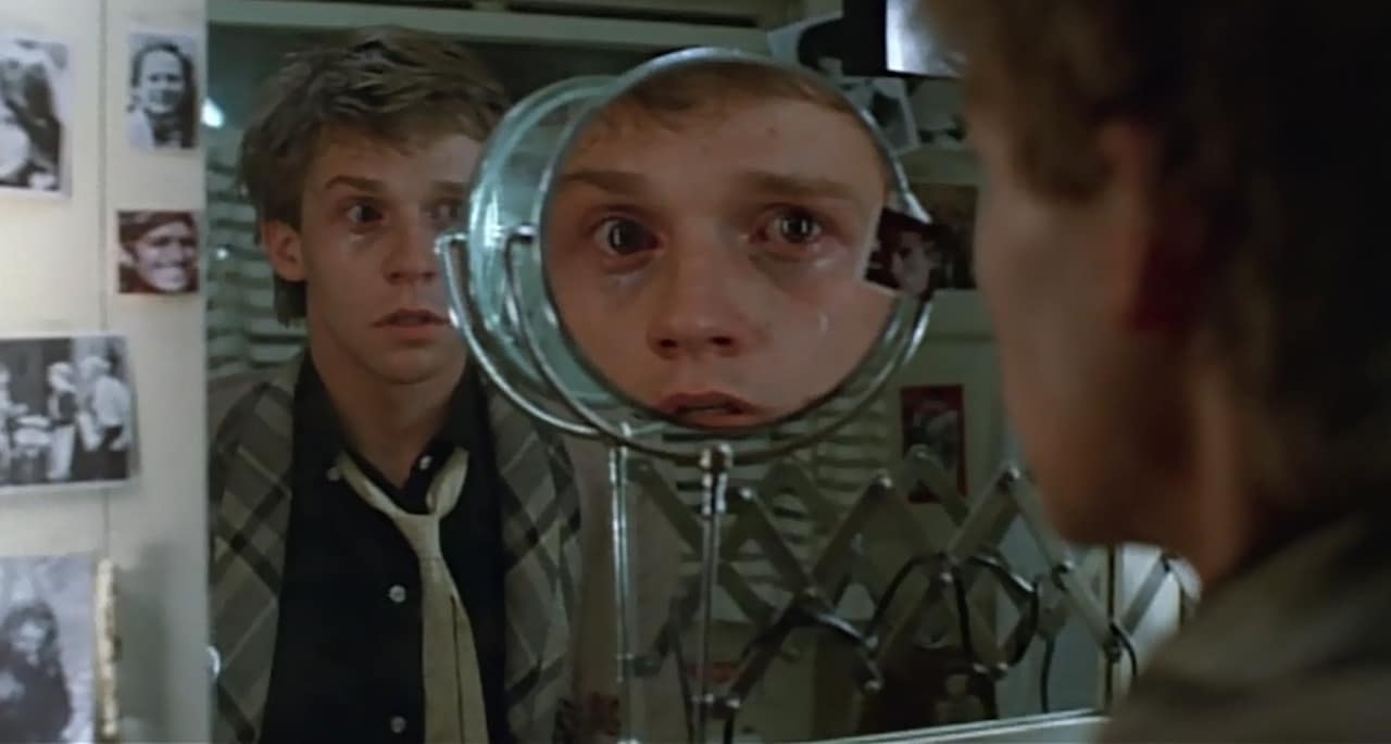 A young man looks at his reflection in a round mirror, which magnifies his face, in a room with black and white photos on the wall.