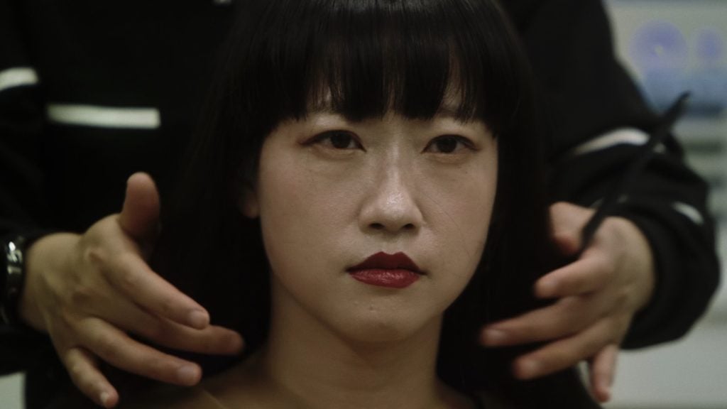 A woman with straight black hair and red lipstick sits still while a person behind her positions their hands near her head, possibly preparing for a haircut.