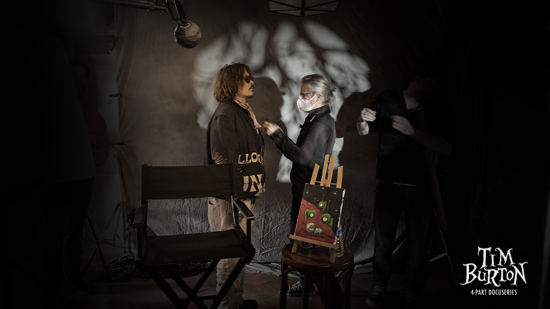 Two people stand facing each other in a dimly lit studio, one adjusting the other's clothing. An easel with a painting and a director's chair are beside them. "Tim Burton" text appears in the corner.