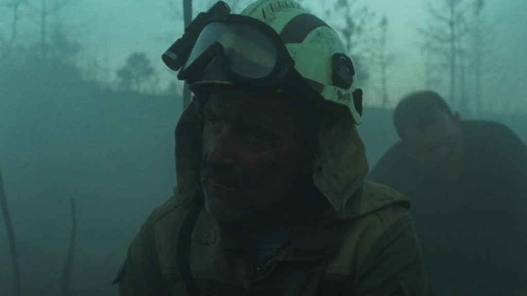 A firefighter wearing a helmet and goggles is covered in soot, standing in a smoky outdoor environment with another figure blurred in the background.
