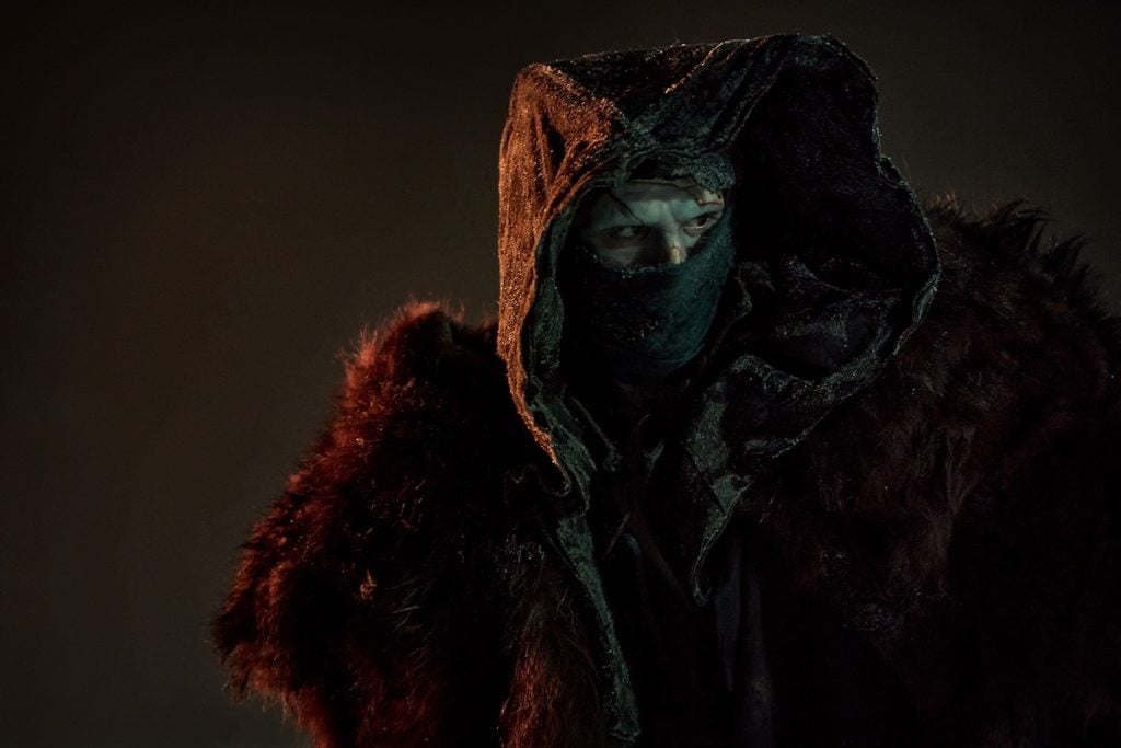 A person wearing a dark hooded cloak, a mask covering the lower half of their face, and a fur garment stands in dim lighting.