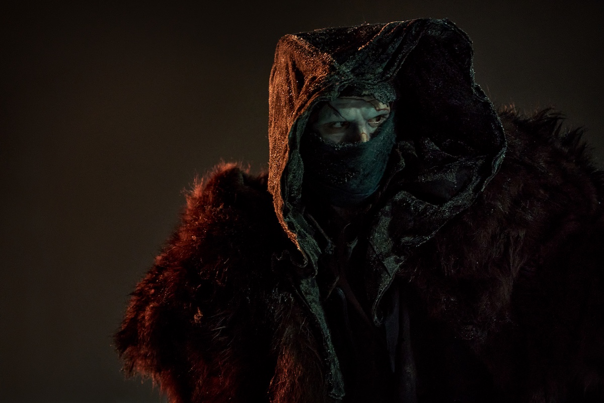 A person wearing a dark hooded cloak, a mask covering the lower half of their face, and a fur garment stands in dim lighting.