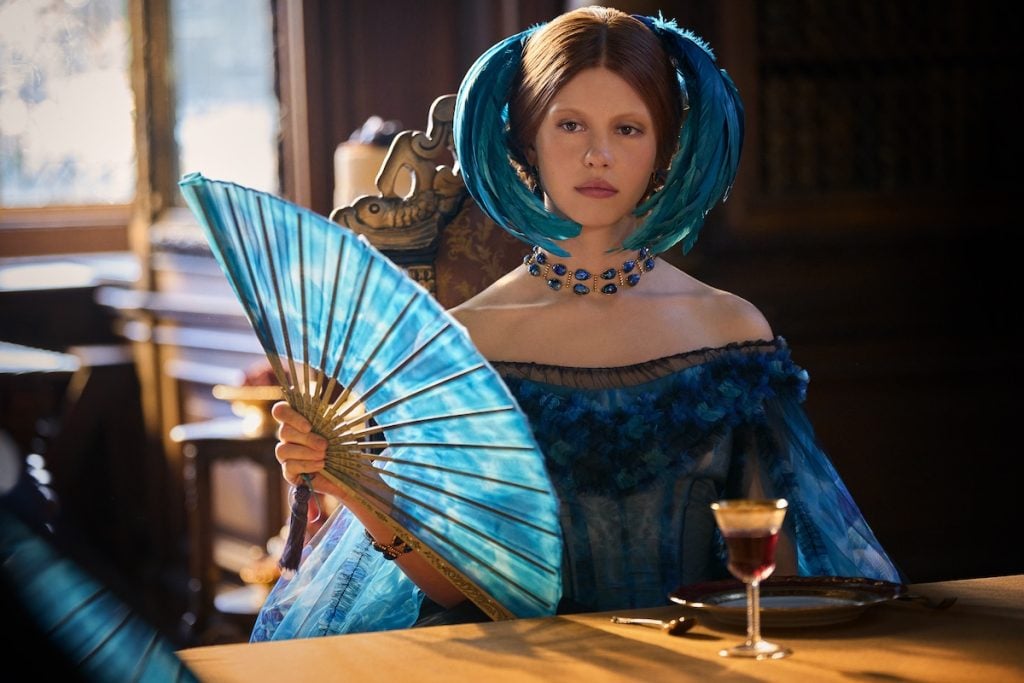 A woman wearing a blue Victorian-style dress and elaborate feathered headpiece sits at a table, holding a large blue fan, with a drink and plate in front of her.