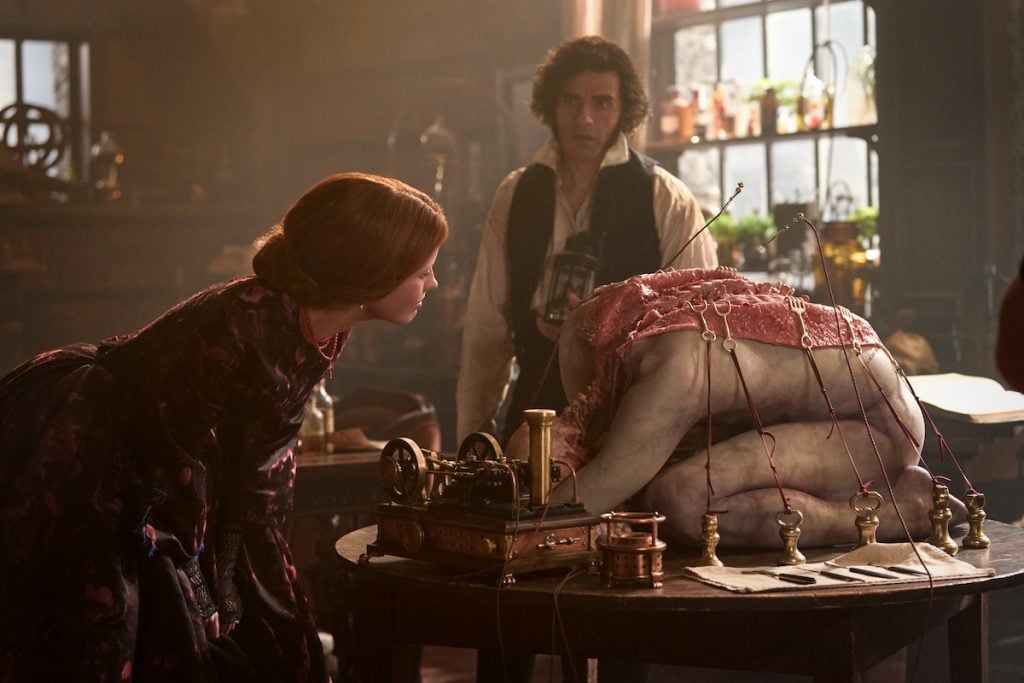 A woman in period clothing inspects a large, human-like figure attached to wires and a machine, while a man stands in the background in a dimly lit room.