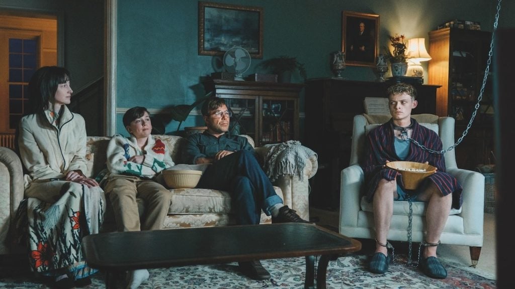 Four people sit in a living room; three on a couch, one in a robe and slippers is chained to a table and holding a bowl. The atmosphere appears tense or unusual.
