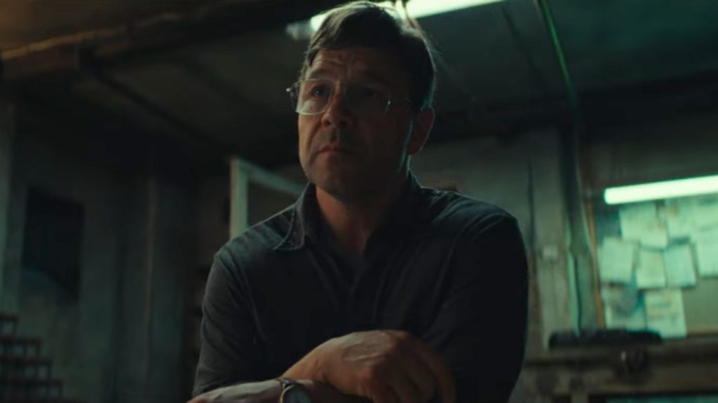 A man with glasses sits in a dimly lit room, crossing his arms and looking slightly upward. Papers are visible on the wall behind him.