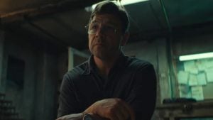 A man with glasses sits in a dimly lit room, crossing his arms and looking slightly upward. Papers are visible on the wall behind him.