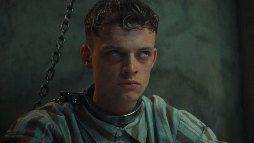 A young man with short curly hair sits against a wall, wearing a striped shirt and a metal collar attached to a chain, looking upward with a serious expression.