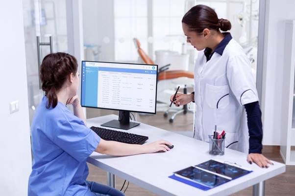Two medical professionals review patient information on a computer in a modern clinic office, with X-ray images and dental equipment visible in the background.