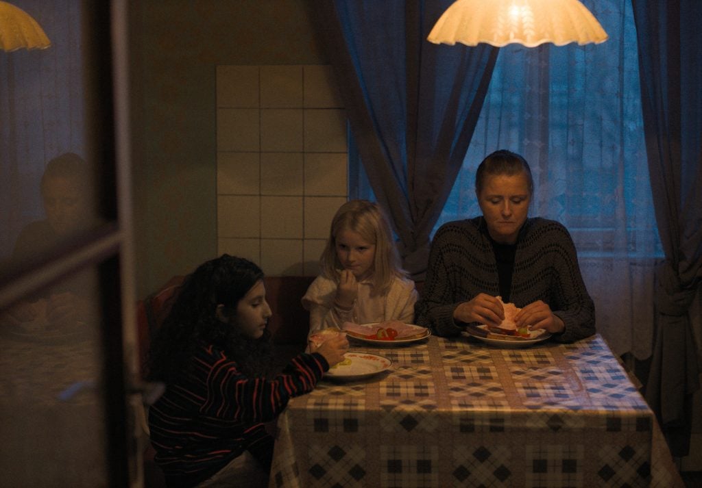Three people sit at a table with food in a dimly lit room; two children sit on the left and an adult on the right, all facing forward under a hanging lamp.
