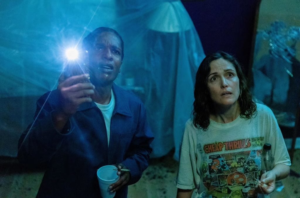 A man holding a flashlight and a cup stands next to a woman in a graphic t-shirt; both look ahead with concerned expressions in a dimly lit room.