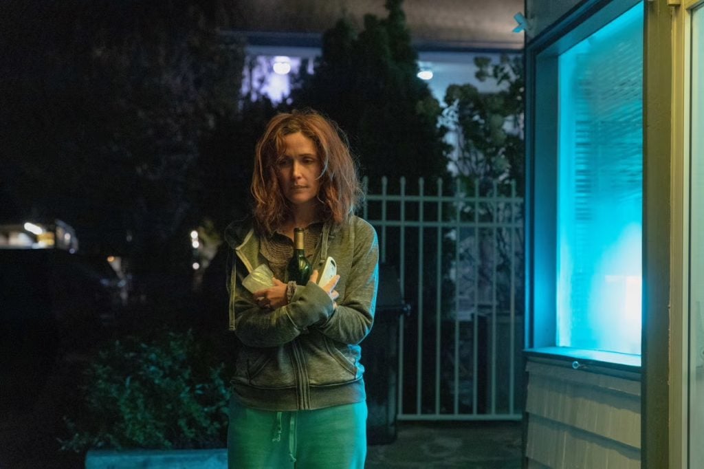 A woman with tousled hair stands outside at night, wearing a grey hoodie and holding several items to her chest, illuminated by blue light from a nearby window.