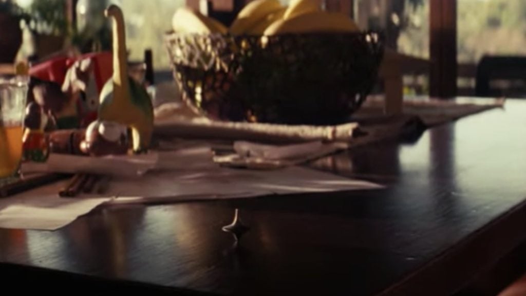 A spinning top on a wooden table in the foreground, with scattered papers and a fruit bowl containing bananas in the background.