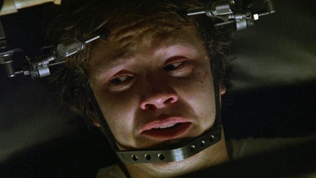 A distressed man with red eyes is restrained by a metal head brace, looking upward with a fearful expression.