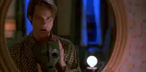 A man stands in front of a mirror, holding a green wooden mask in one hand and wearing a patterned robe. His mouth is open and a lit bulb is visible in the foreground.