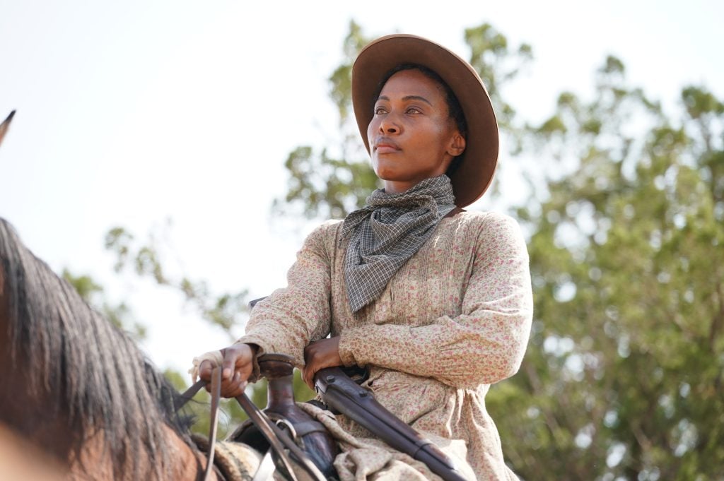 A woman wearing a wide-brimmed hat and patterned dress rides a horse outdoors, holding the reins and looking ahead with a serious expression. Trees are visible in the background.